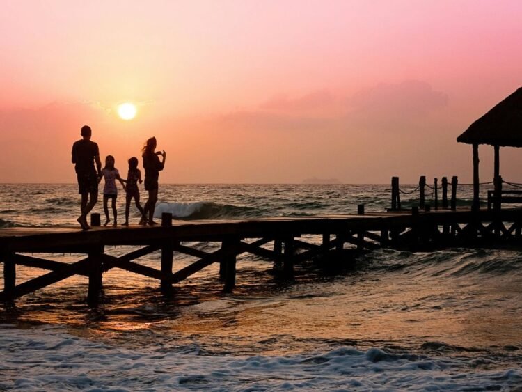 Na imagem vemos uma família aproveitando as férias caminhando por uma passarela de madeira sobre o mare ao fundo um lindo por sol, mostrando que é possível fazer uma viagem econômica em família