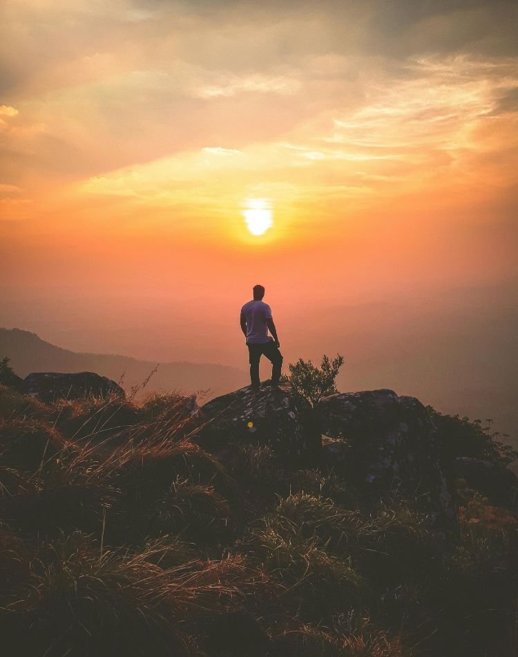 Na foto vemos uma pessoa que está viajando sozinha em pé no alto de uma montanha assistindo a um lindo por do sol alaranjado. essa imagem demonstra como alguém viajando sozinho sem erros pode curtir a natureza.