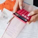 Uma pessoa segura uma calculadora rosa enquanto faz anotações e cálculos com um lápis. Sobre a mesa estão espalhados diversos comprovantes, notas fiscais e papéis com dados financeiros. A cena representa o momento de organização financeira, ideal para ilustrar temas como orçamento familiar, controle de gastos, finanças pessoais ou planejamento econômico.
