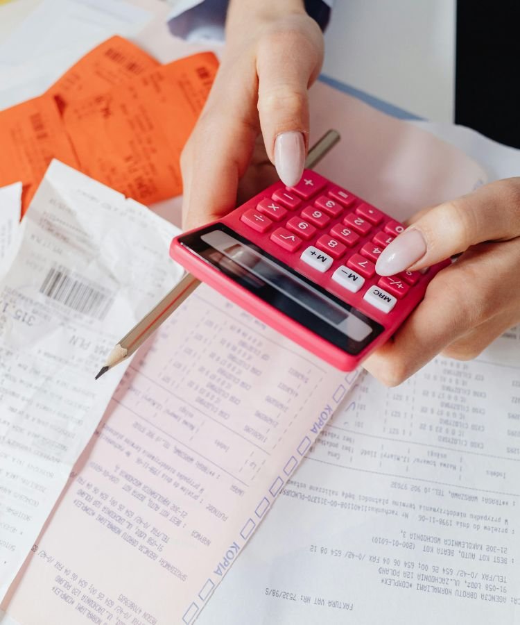 Uma pessoa segura uma calculadora rosa enquanto faz anotações e cálculos com um lápis. Sobre a mesa estão espalhados diversos comprovantes, notas fiscais e papéis com dados financeiros. A cena representa o momento de organização financeira, ideal para ilustrar temas como orçamento familiar, controle de gastos, finanças pessoais ou planejamento econômico.