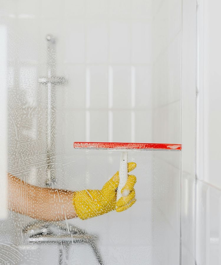 Pessoa com luva amarela limpando o box do banheiro com rodo de limpeza. A imagem ilustra uma das tarefas práticas do cronograma de limpeza para quem tem pouco tempo, ideal para manter a casa em ordem mesmo com uma rotina corrida.