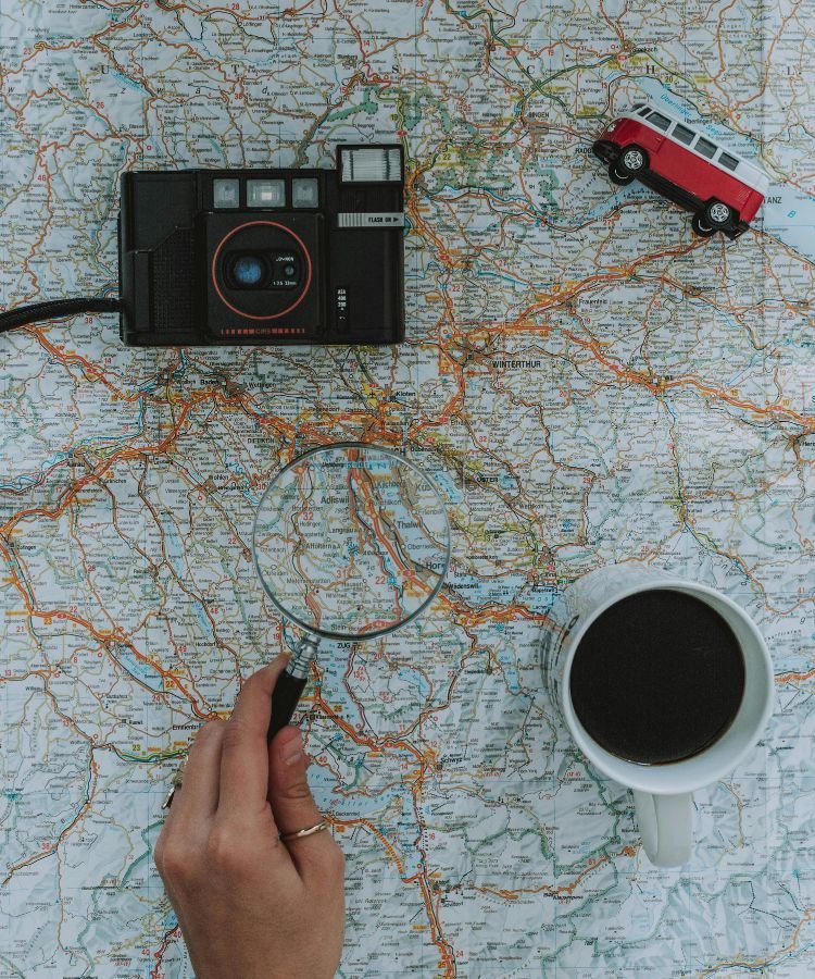 Na imagem, vemos uma representação perfeita do espírito aventureiro e da preparação essencial para uma viagem inesquecível. Um mapa detalhado serve de fundo para itens simbólicos: uma câmera vintage, uma miniatura de kombi vermelha, uma xícara de café quente e uma lupa segurada por uma mão curiosa — todos elementos que evocam a descoberta, o planejamento e o entusiasmo por explorar novos destinos. Essa composição transmite visualmente como um bom roteiro de viagem pode transformar a experiência, trazendo organização, economia de tempo e liberdade para aproveitar cada momento ao máximo.